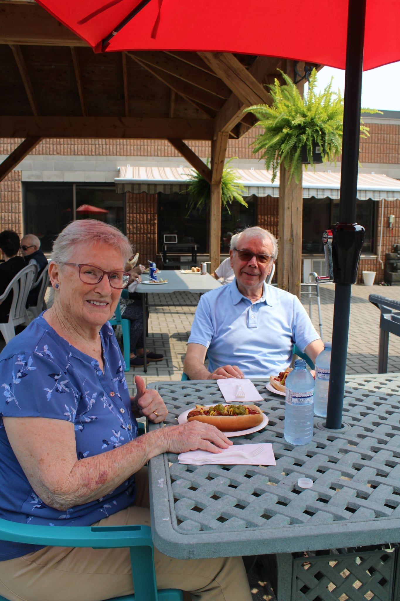 Two people sitting at an outdoor café table under a red umbrella, with drinks and food.