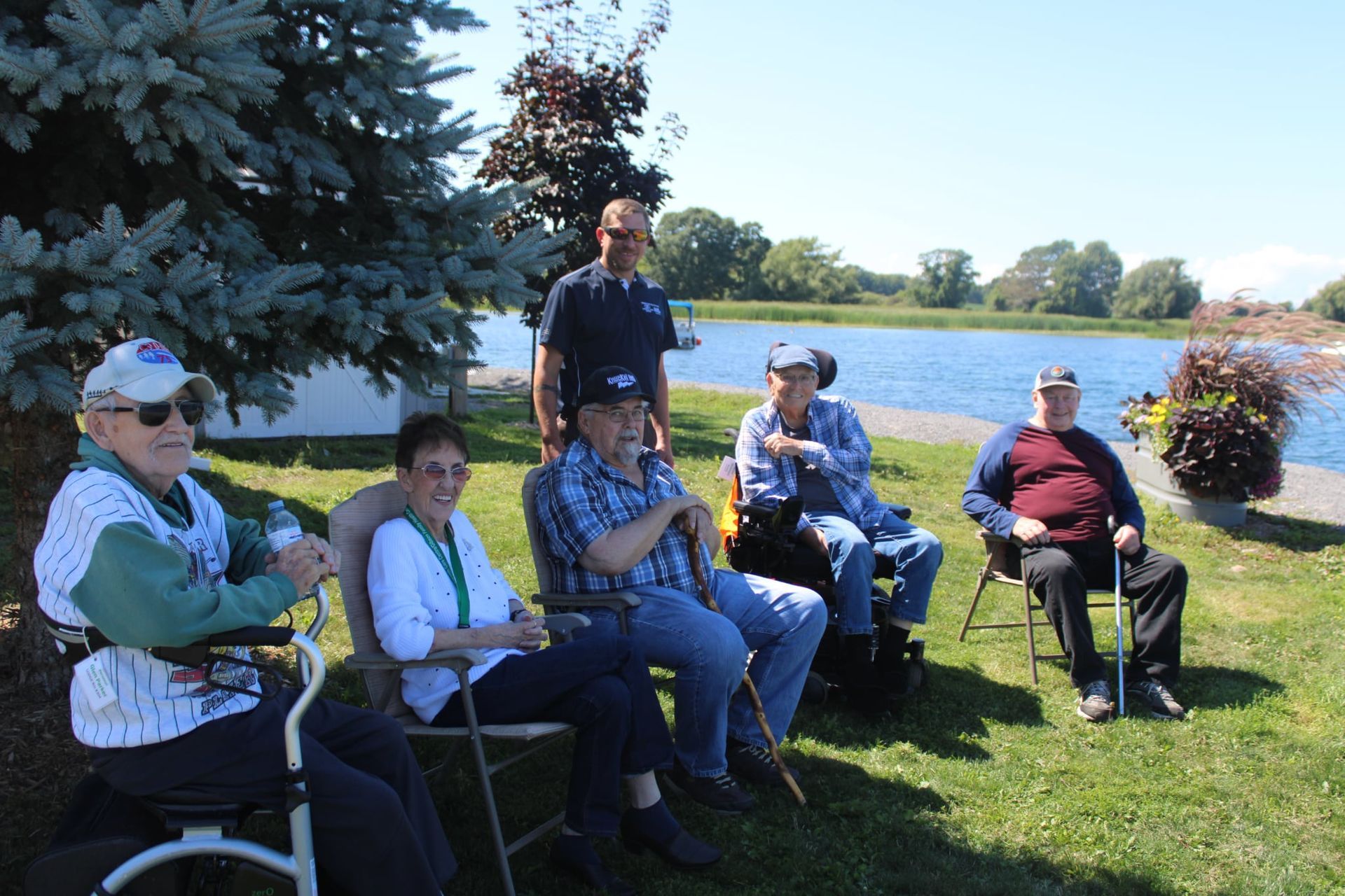 People seated outdoors on folding chairs in a grassy park, with a man standing behind them near a tree.