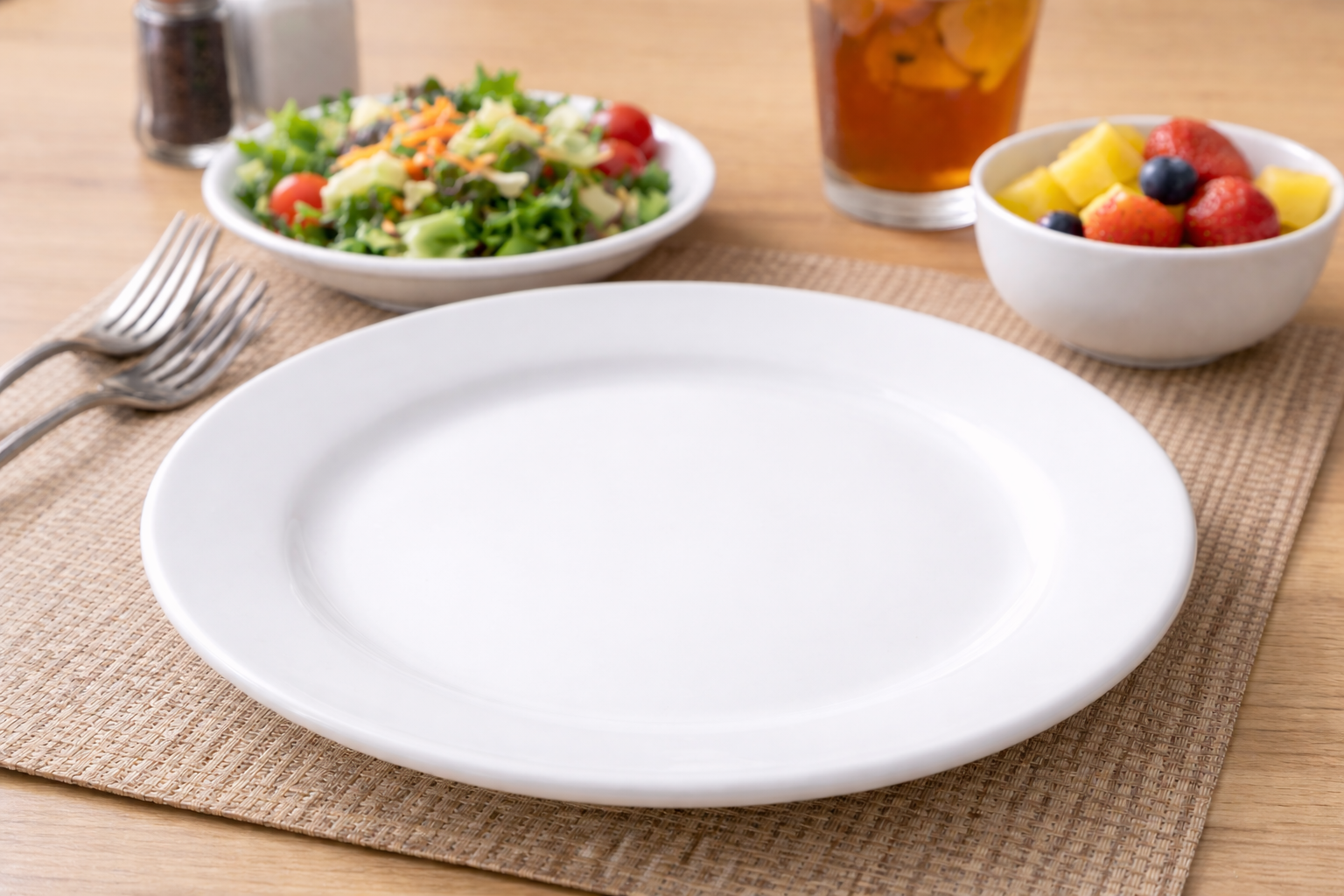 Empty white plate on a woven placemat with salad, fruit, utensils, and drinks on a table