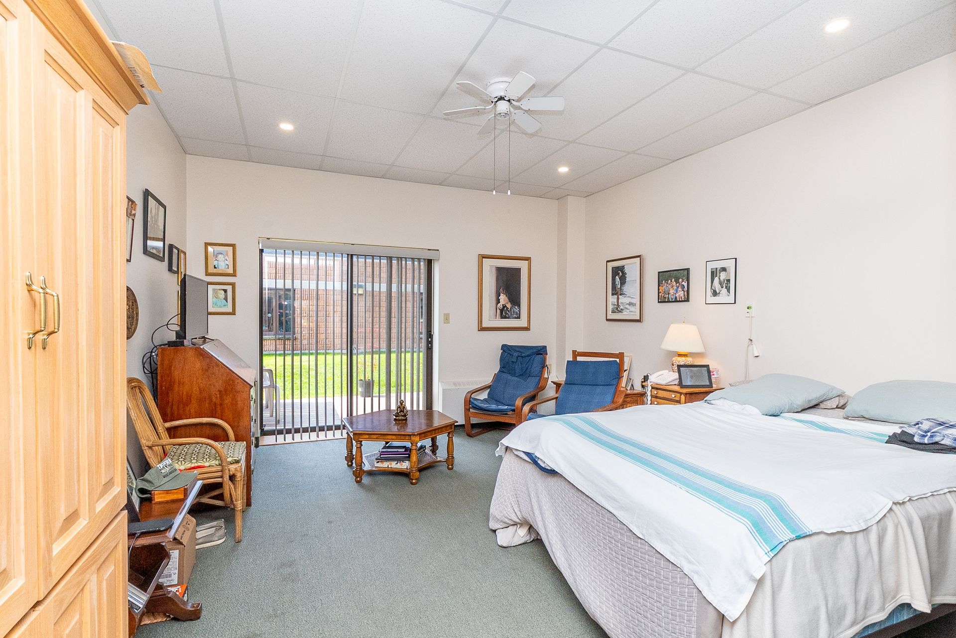 Spacious bedroom with a bed, wicker chair, blue armchairs, framed art, and a sliding glass door to a patio.