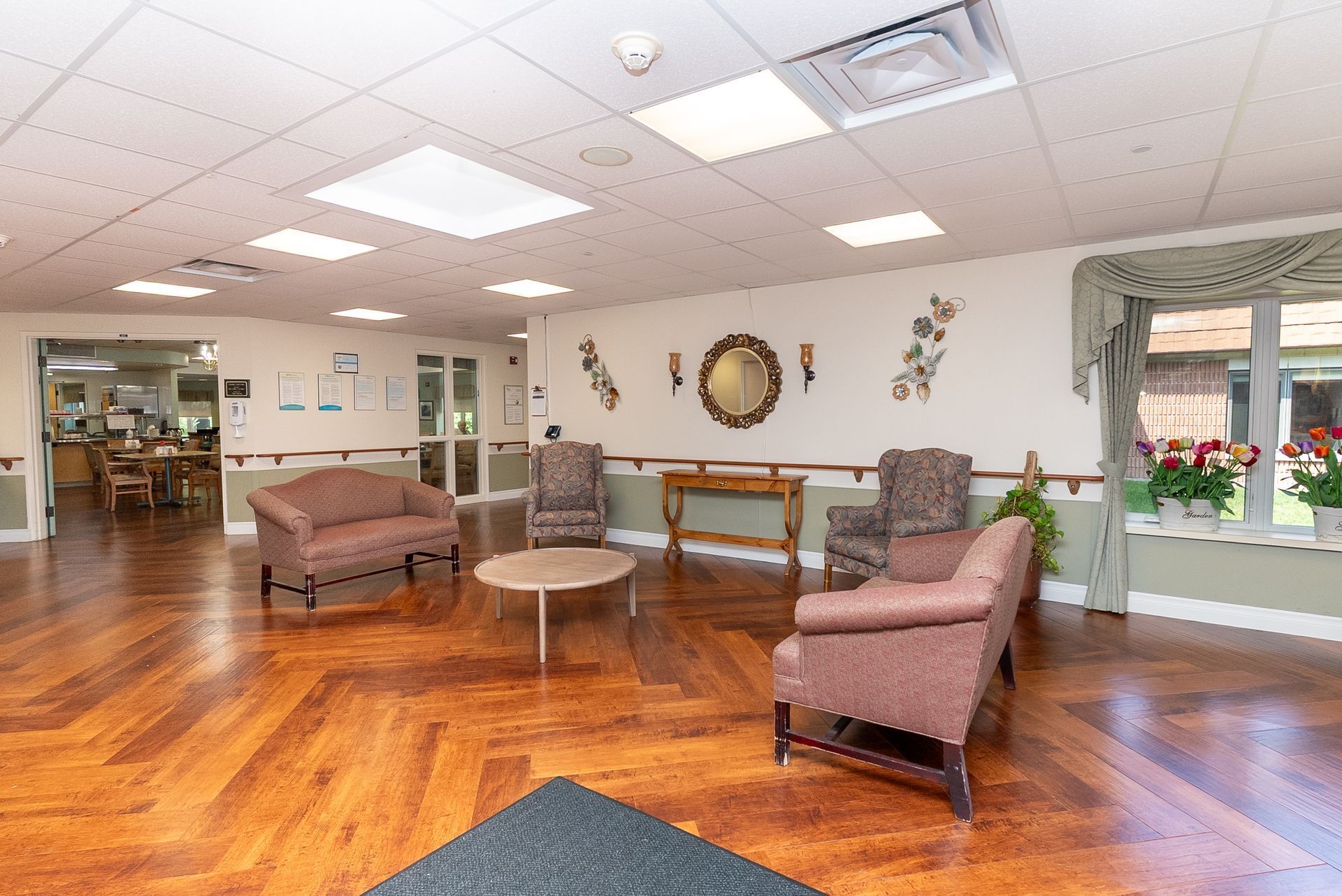 Bright lobby with hardwood floors, pink chairs, a table, and large windows with plants.