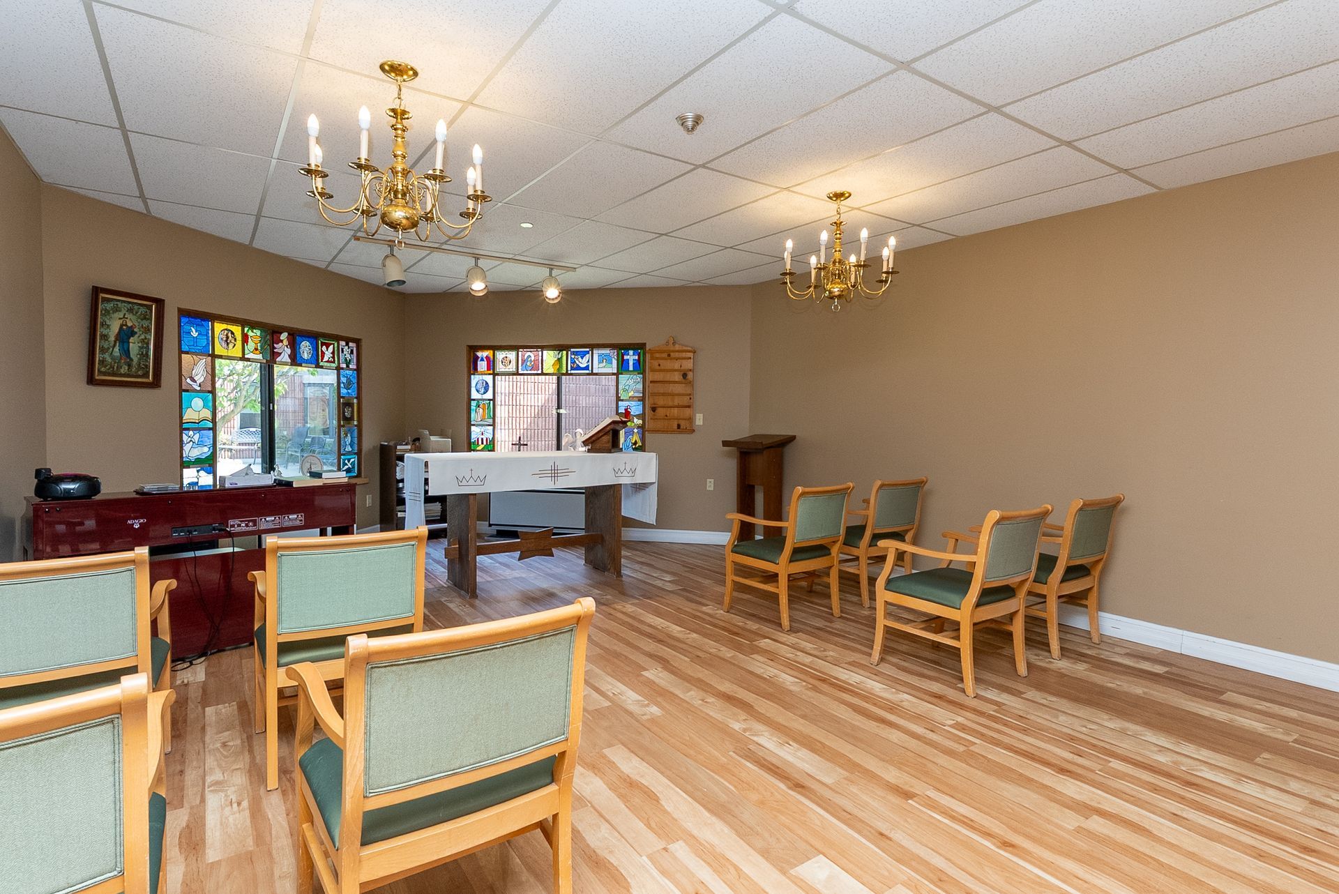 Empty community room with chairs, chandeliers, polished wood floor, and a small stage at the back.