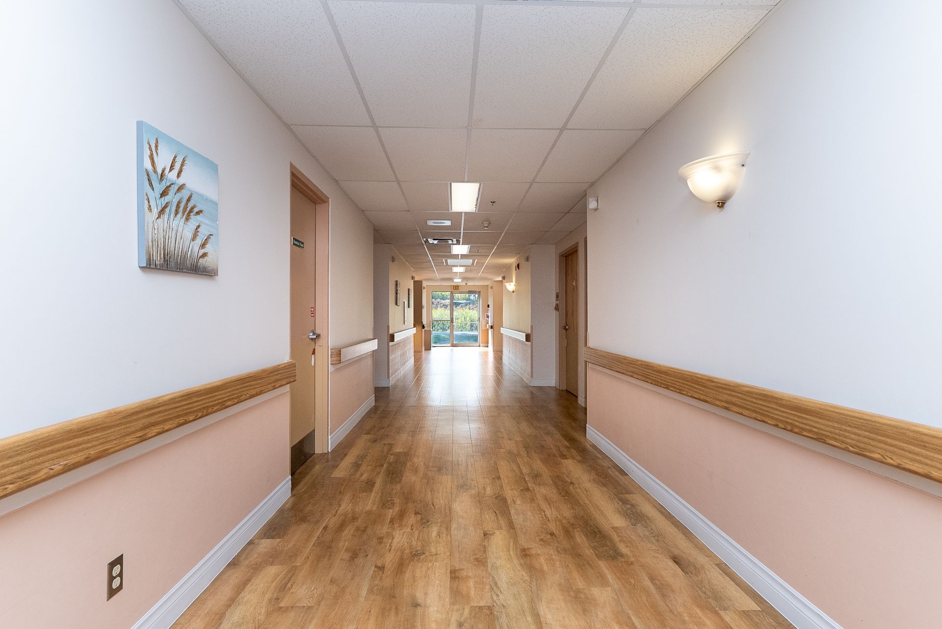 Bright hospital hallway with beige walls, wood trim, and fluorescent ceiling lights leading to a distant doorway