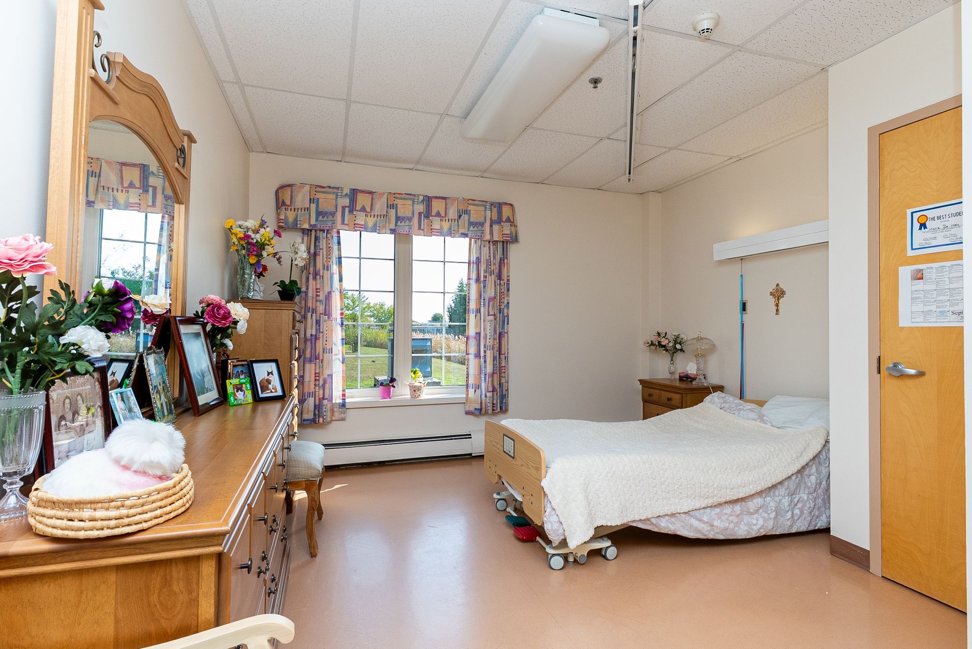 Small bedroom with a single bed, dresser with flowers, and window with curtains and a view outside