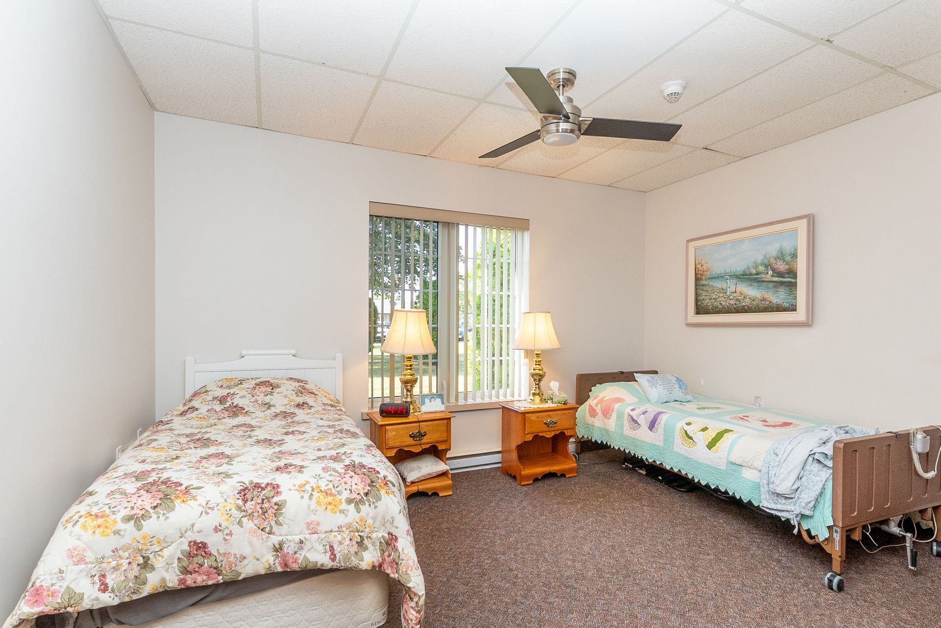 Bedroom with two twin beds, lamp tables, window blinds, ceiling fan, and framed wall art.