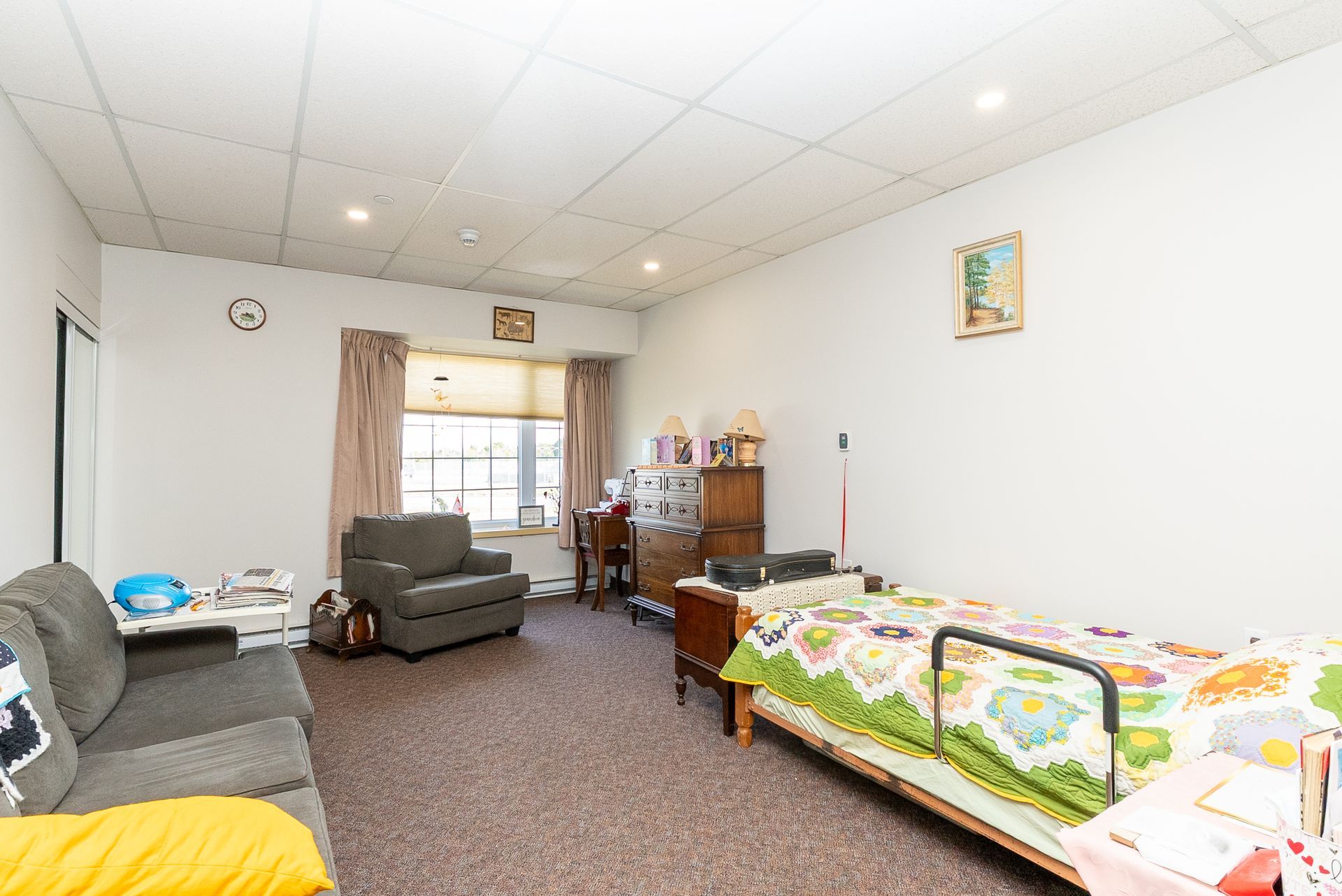 Bright living room with gray sofas, a recliner, and a daybed with a colorful quilt near a window.