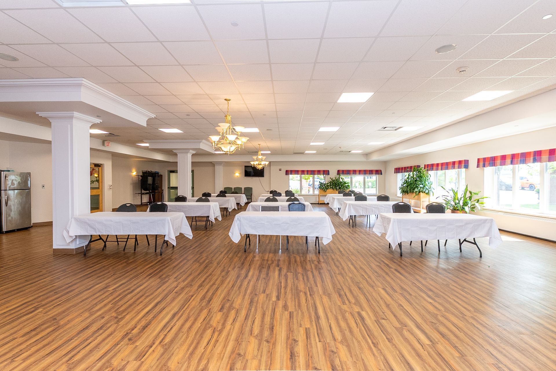Spacious banquet hall with wooden floors, white tablecloth tables, chandeliers, and large windows