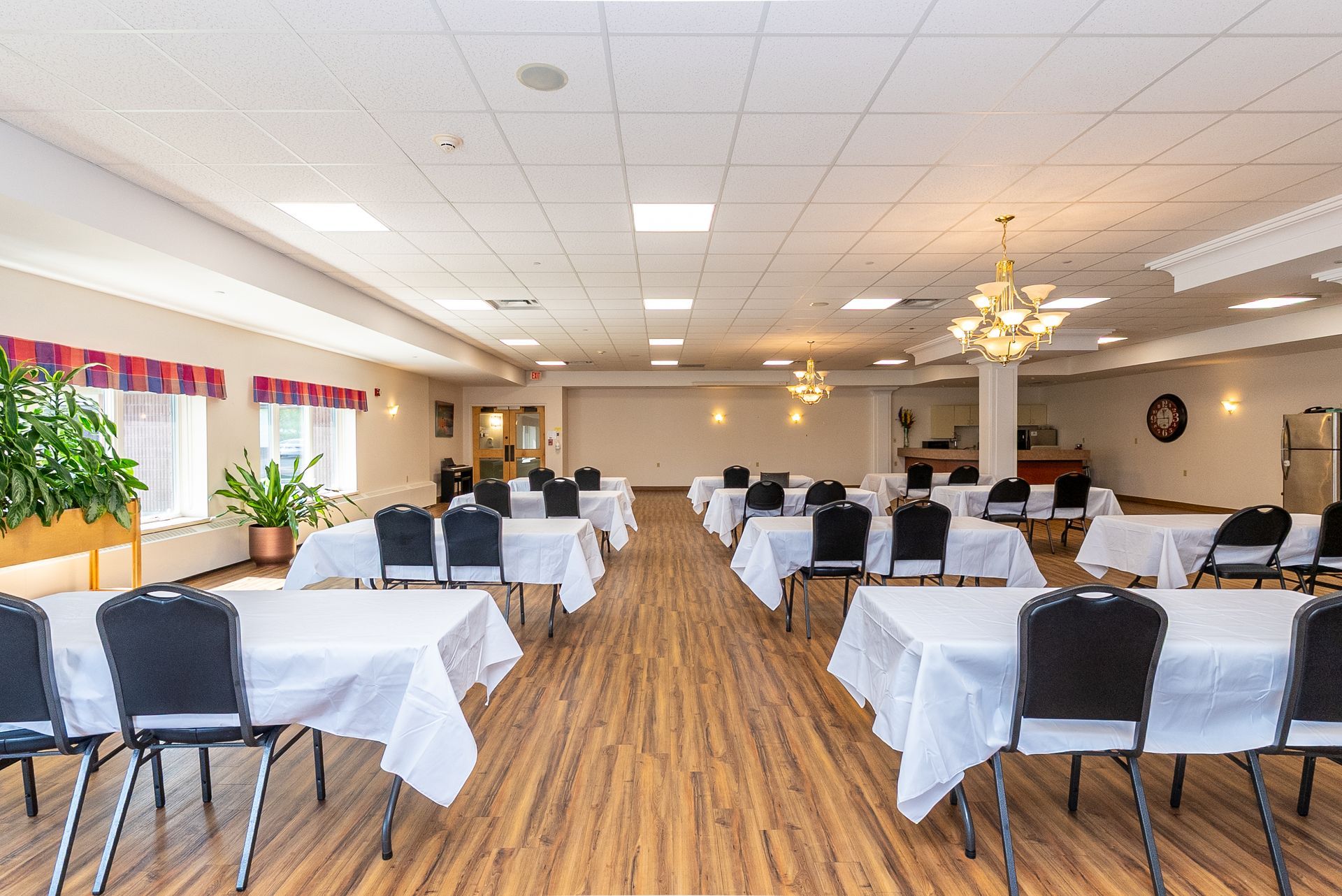 Dining hall with white tablecloths, black chairs, wood floor, and ceiling lights in a bright banquet room