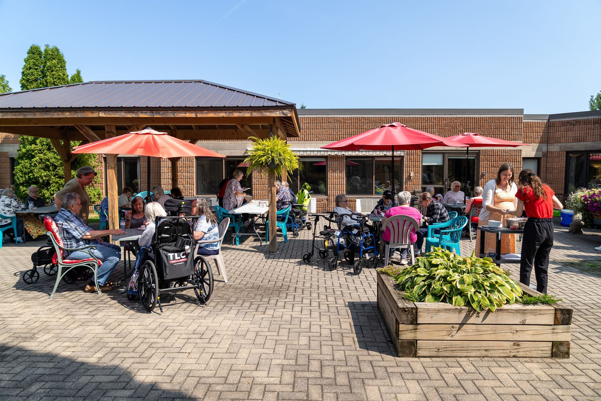 Outdoor café patio with red umbrellas, people seated at tables, and a few strollers on a sunny day
