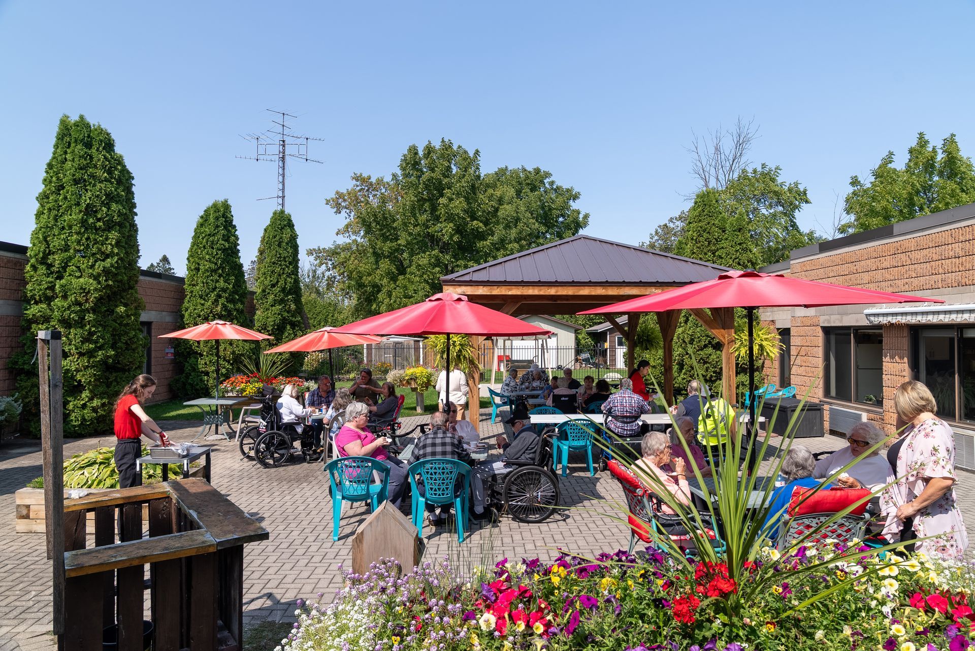 Outdoor café patio with red umbrellas, seated diners, and colorful flowers on a sunny day