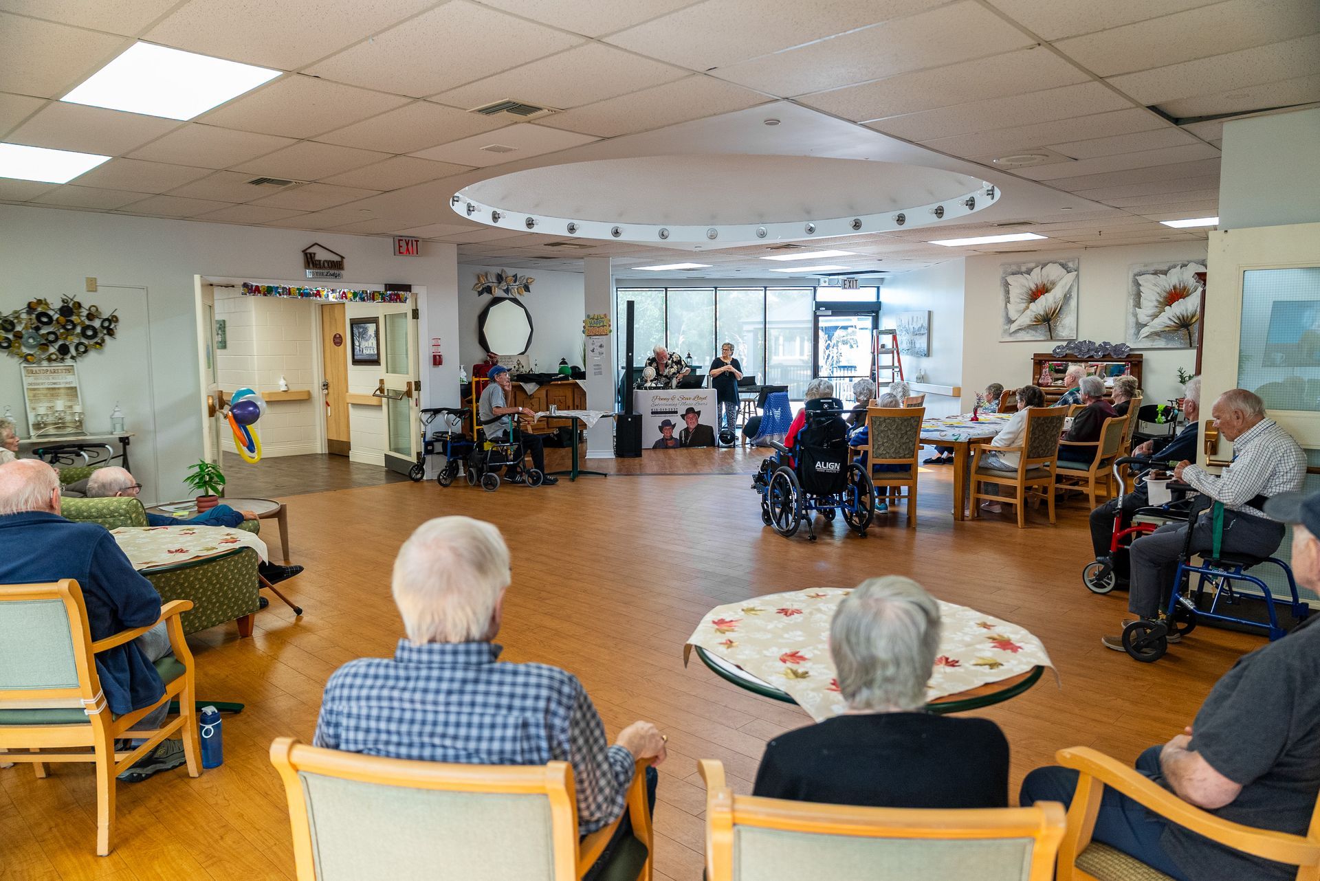 Elder care common room with several people seated in chairs and wheelchairs, facing a bright open lounge.