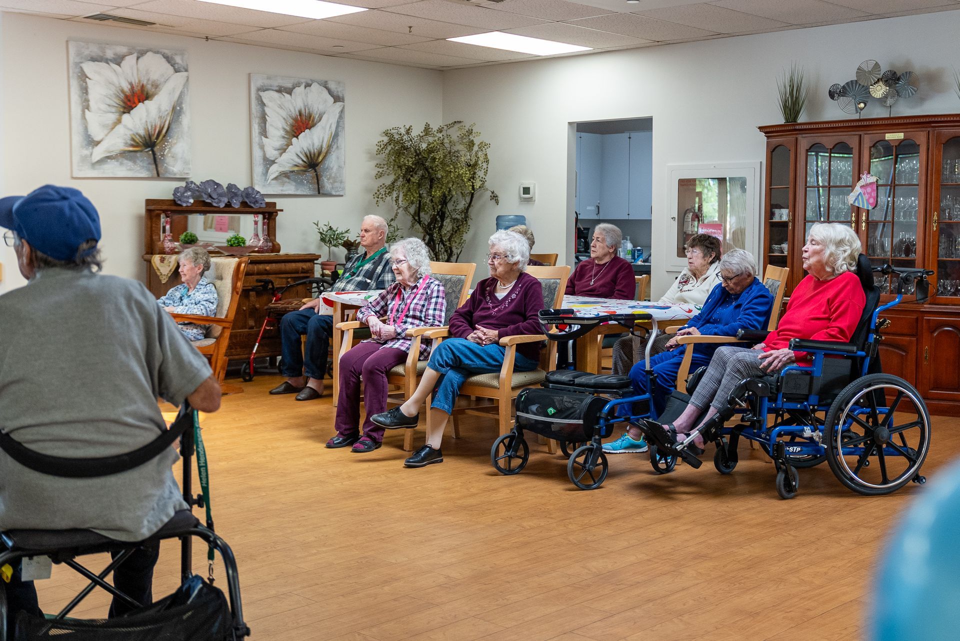 Seniors seated in a community room during an activity, with wheelchairs, armchairs, and floral wall art.