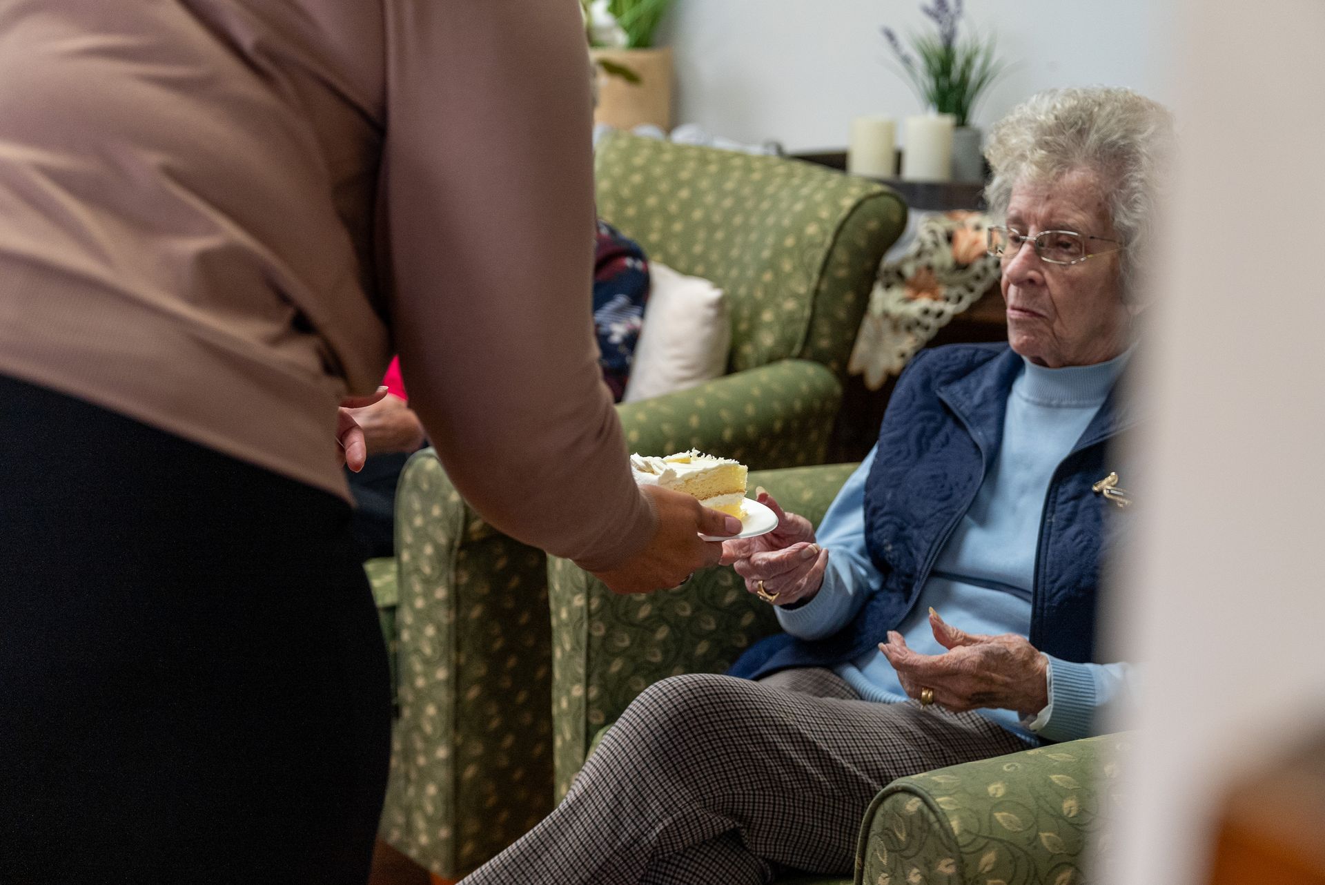 Caregiver handing a drink to an elderly woman seated in a green armchair in a living room