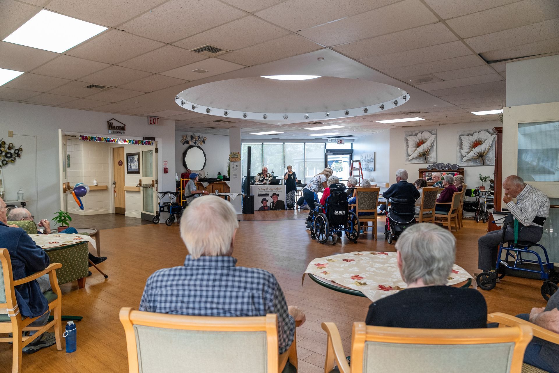 Residents seated in a bright communal room during a group activity, with chairs and tables facing a TV.