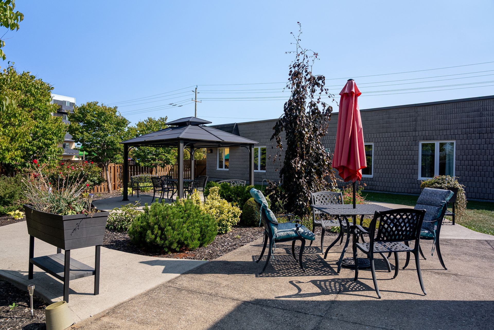 Outdoor patio with tables, chairs, red umbrella, and landscaped garden beside a building