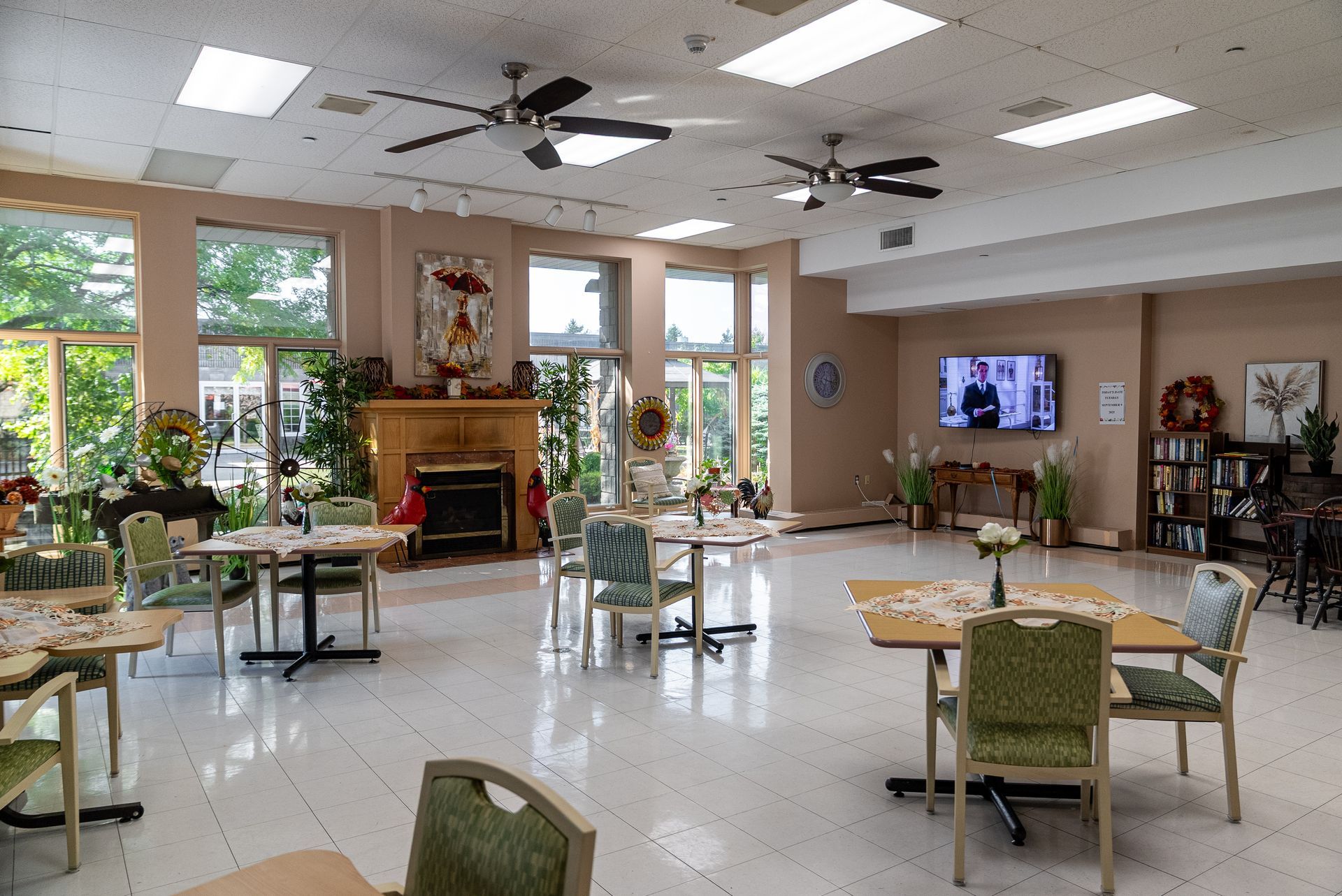 Bright, spacious lounge with tables, green chairs, fireplace, windows, and a wall-mounted TV.