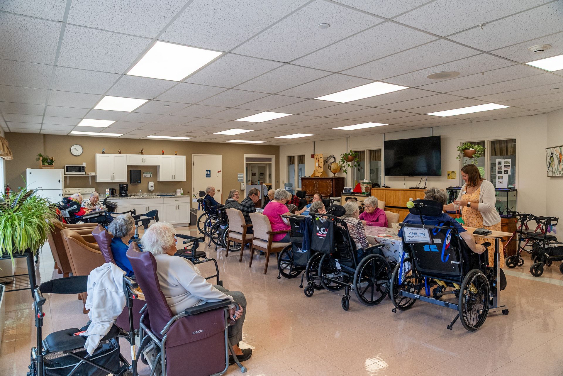 People in wheelchairs gathered in a bright community room, socializing around tables.