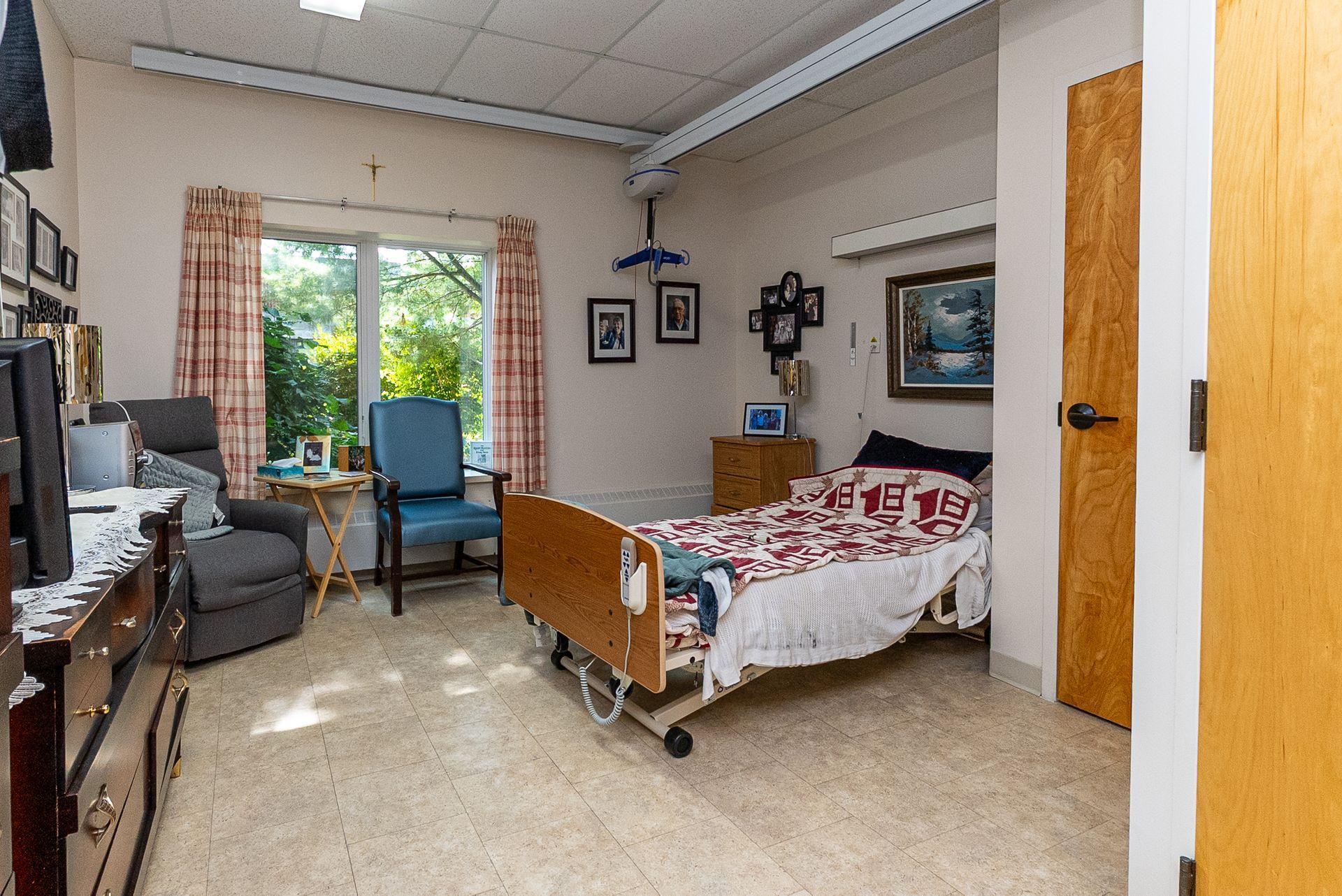 Hospital room with a bed, armchair, TV, and a window overlooking greenery.