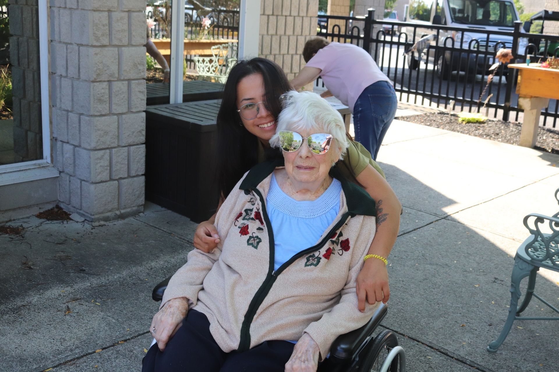 Older woman in a wheelchair outdoors, smiling while another person stands behind her near a building.