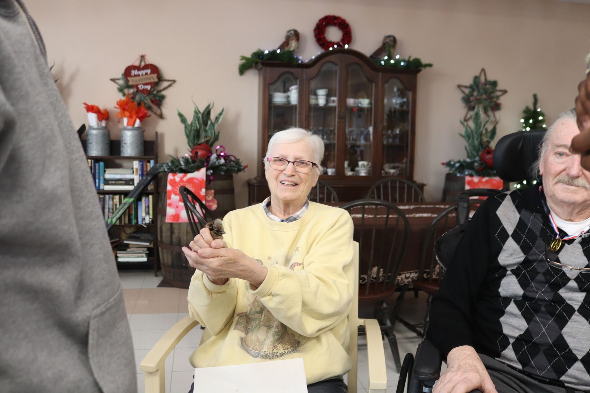 Smiling woman in a yellow sweater clapping in a decorated living room beside another seated person