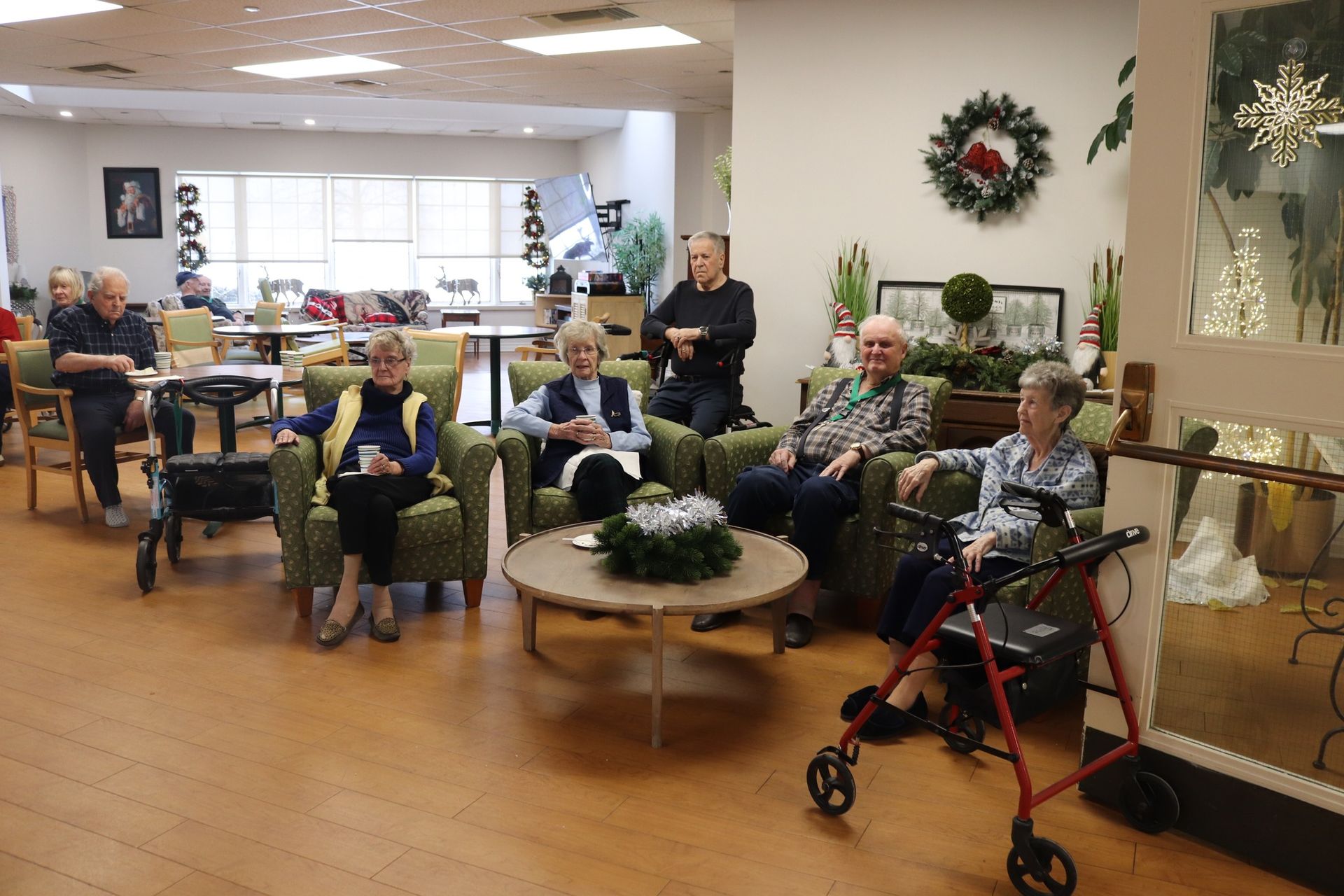 Residents seated in a bright lounge, some using walkers and wheelchairs, with holiday wreaths and decorations
