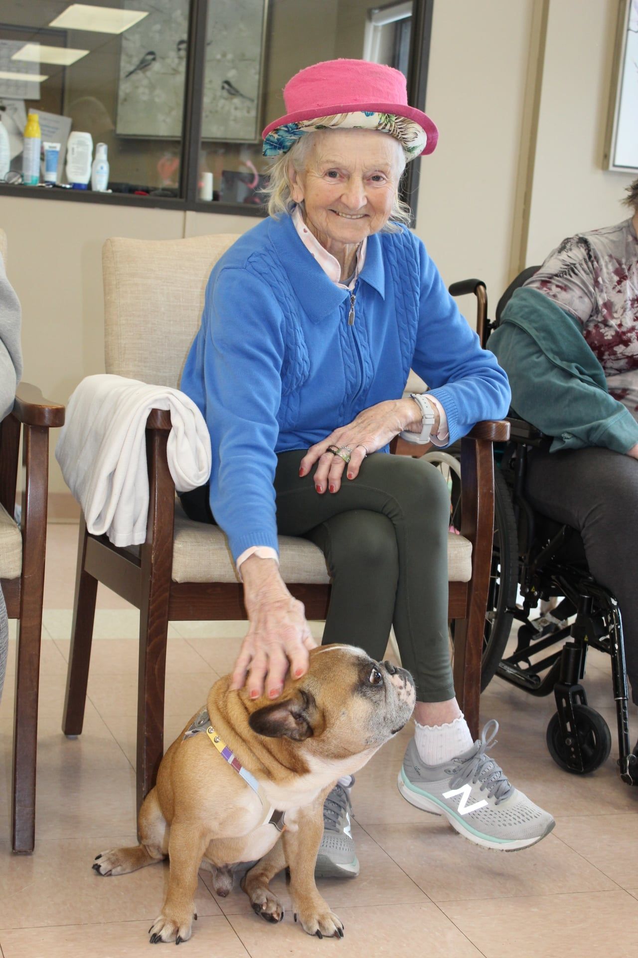Woman in pink hat sits with a bulldog in a room, smiling and petting the dog.