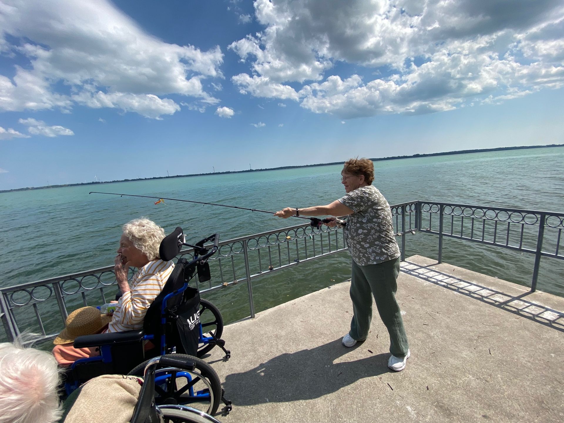 Person pushing a wheelchair beside a waterfront railing under a cloudy blue sky.