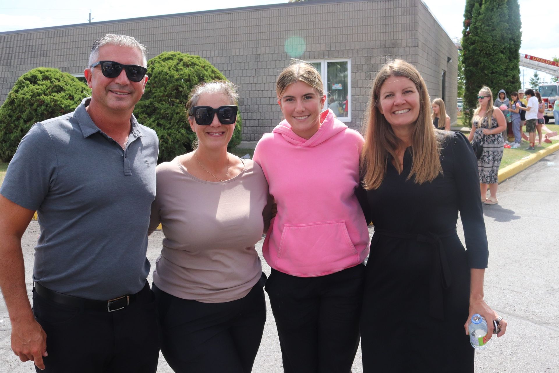 Four people standing outdoors, smiling, with a brick building and people in the background.