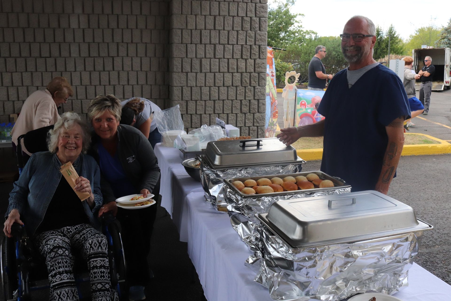 Outdoor buffet table with covered trays and desserts, with people serving themselves near a brick wall.