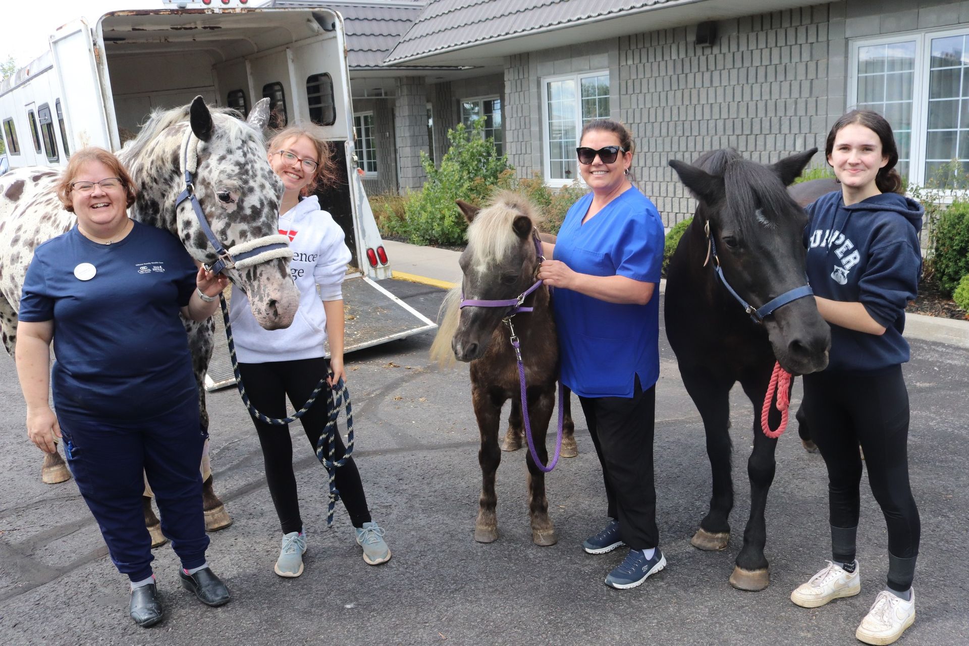 Four people stand with three horses beside an open trailer in a driveway.