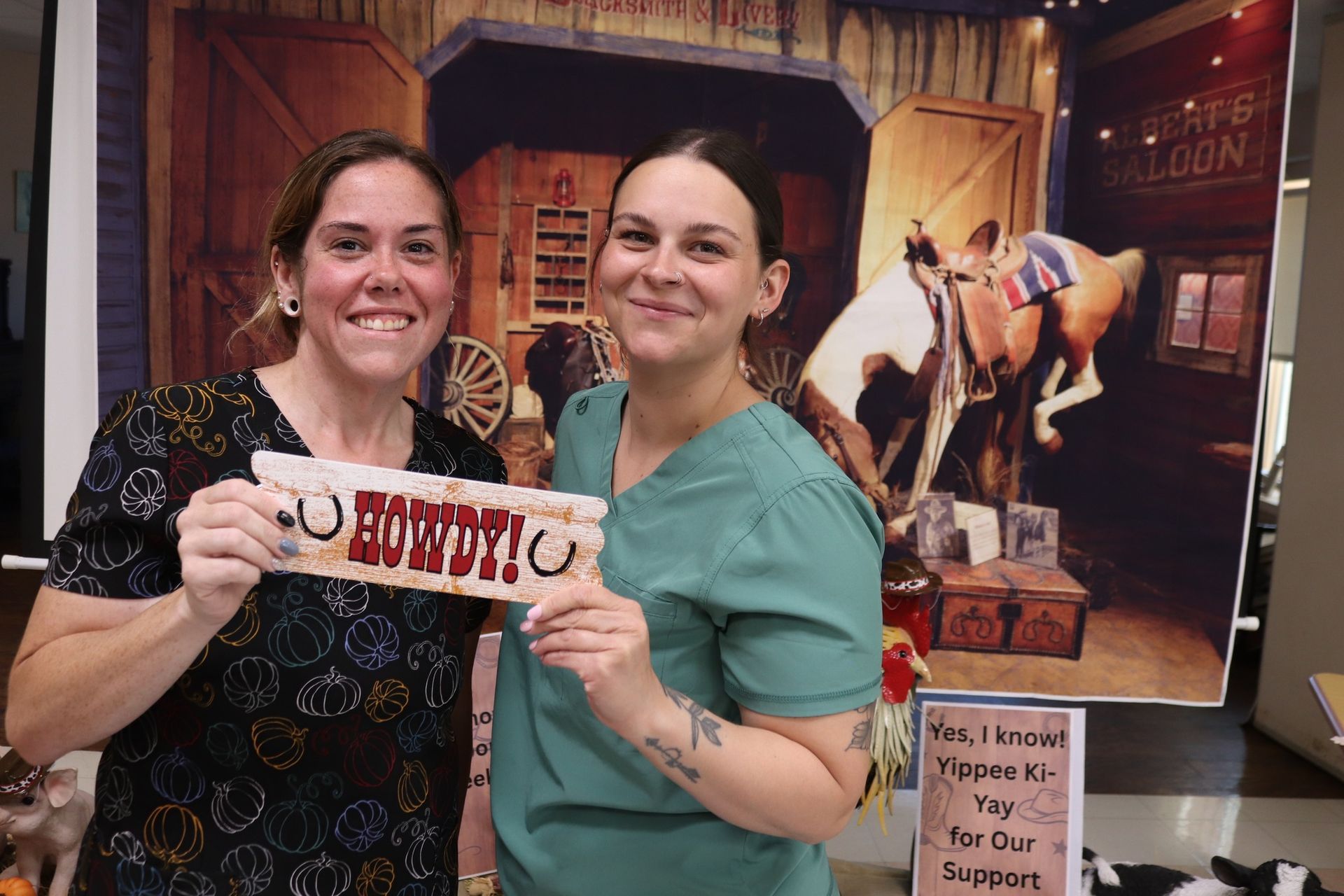 Two women holding a “Howdy” sign at a rustic Western-themed event booth.