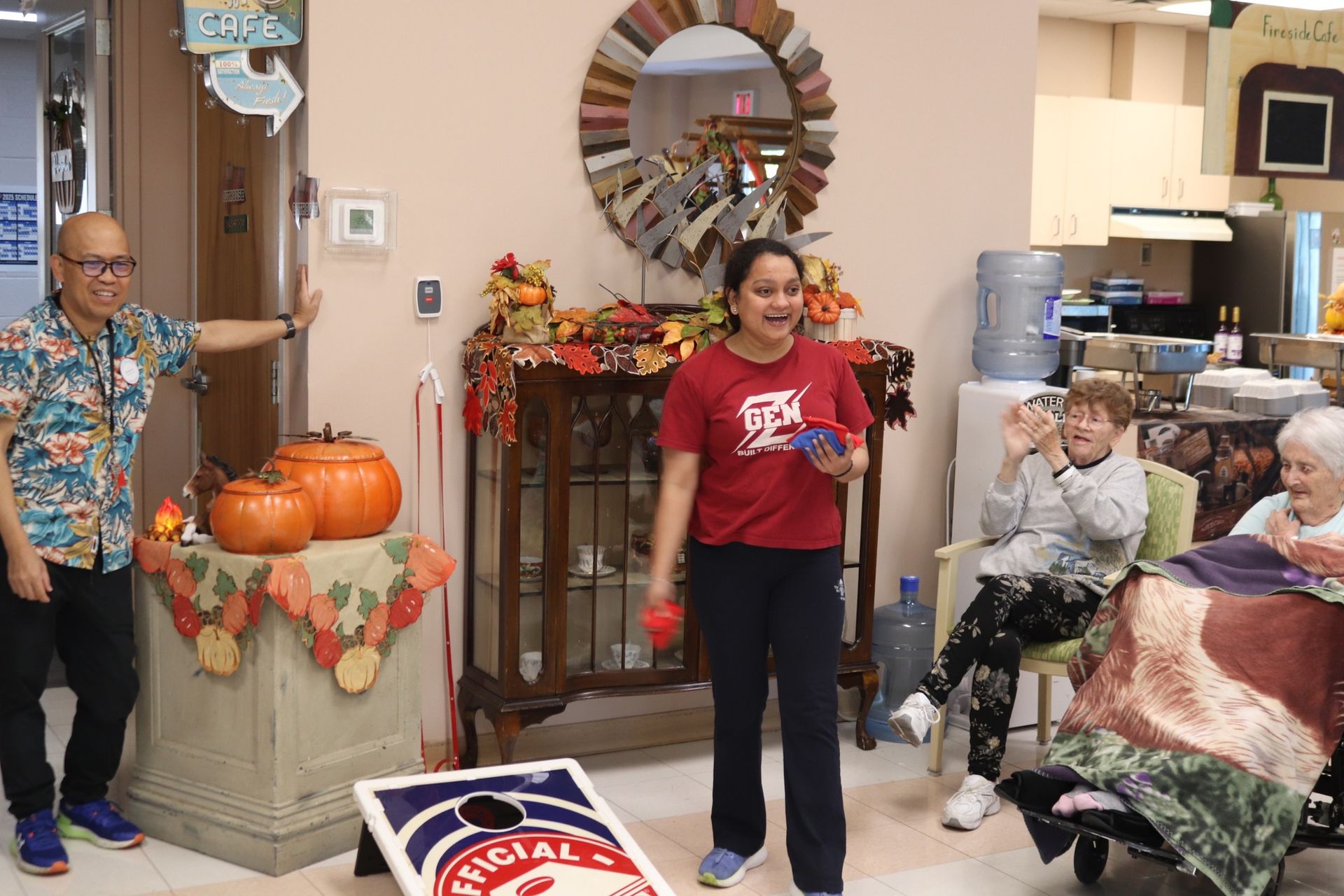 People in a decorated room, with pumpkins by a doorway and seated residents watching a young woman hold red flowers.