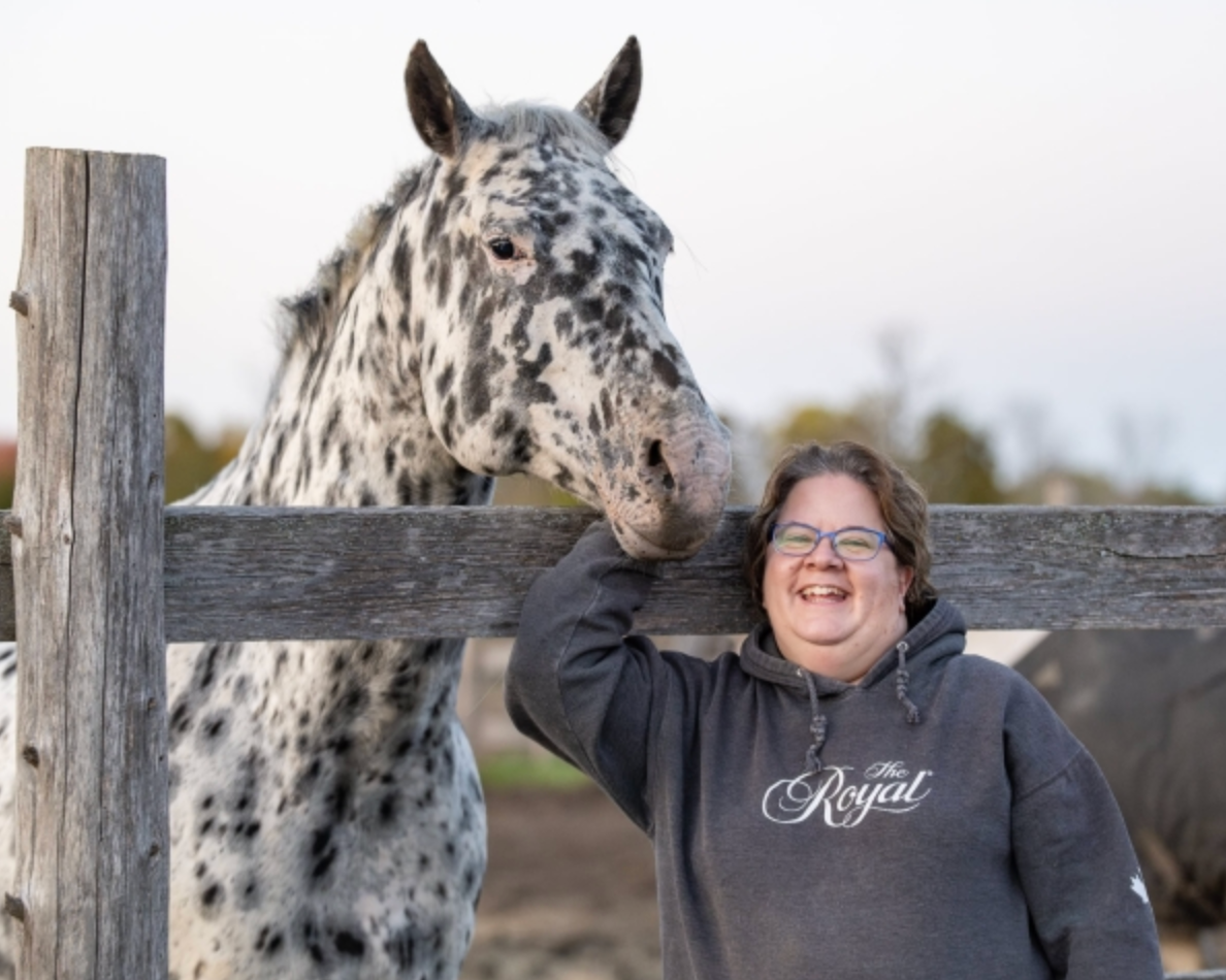 Woman in a dark hoodie smiling beside a dappled horse leaning over a fence.