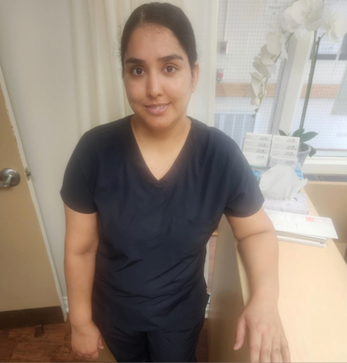Person in dark navy scrubs standing in a clinic room beside a desk and window.