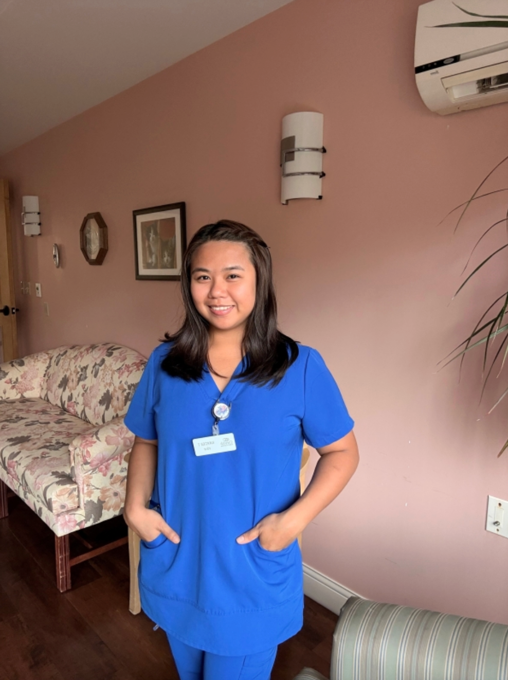 Person in blue scrubs smiling in a pink-walled room with floral furniture and wall decor