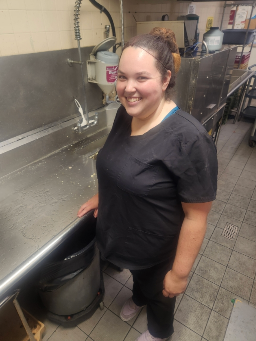 Smiling person in black shirt standing at a stainless steel sink in a kitchen.