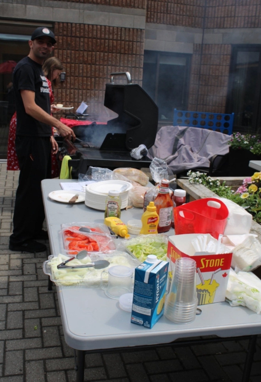 Man grilling outdoors beside a table with food, plates, drinks, and condiments set up for a cookout