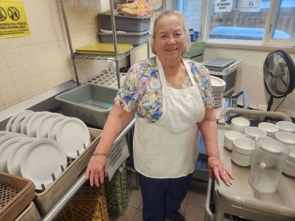 Person in a kitchen apron standing by a sink with stacked dishes and glasses, smiling.
