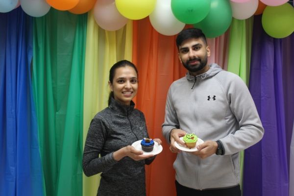 Two people holding small bowls in front of a rainbow balloon and curtain backdrop