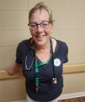 Smiling healthcare worker in navy top with stethoscope, standing against a beige wall.