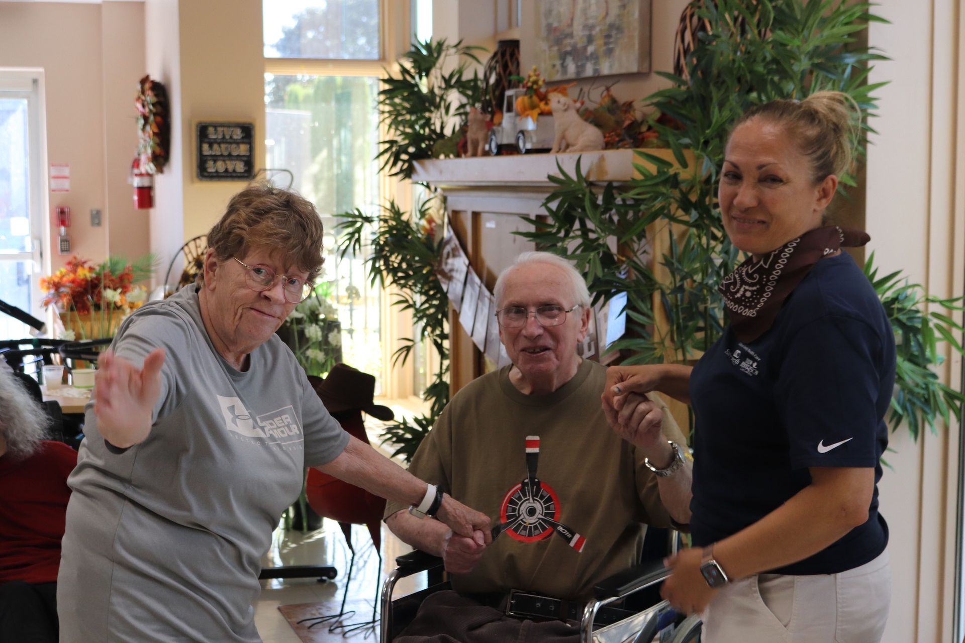 Three people posing indoors, smiling and waving in a bright living room with plants and decorations.