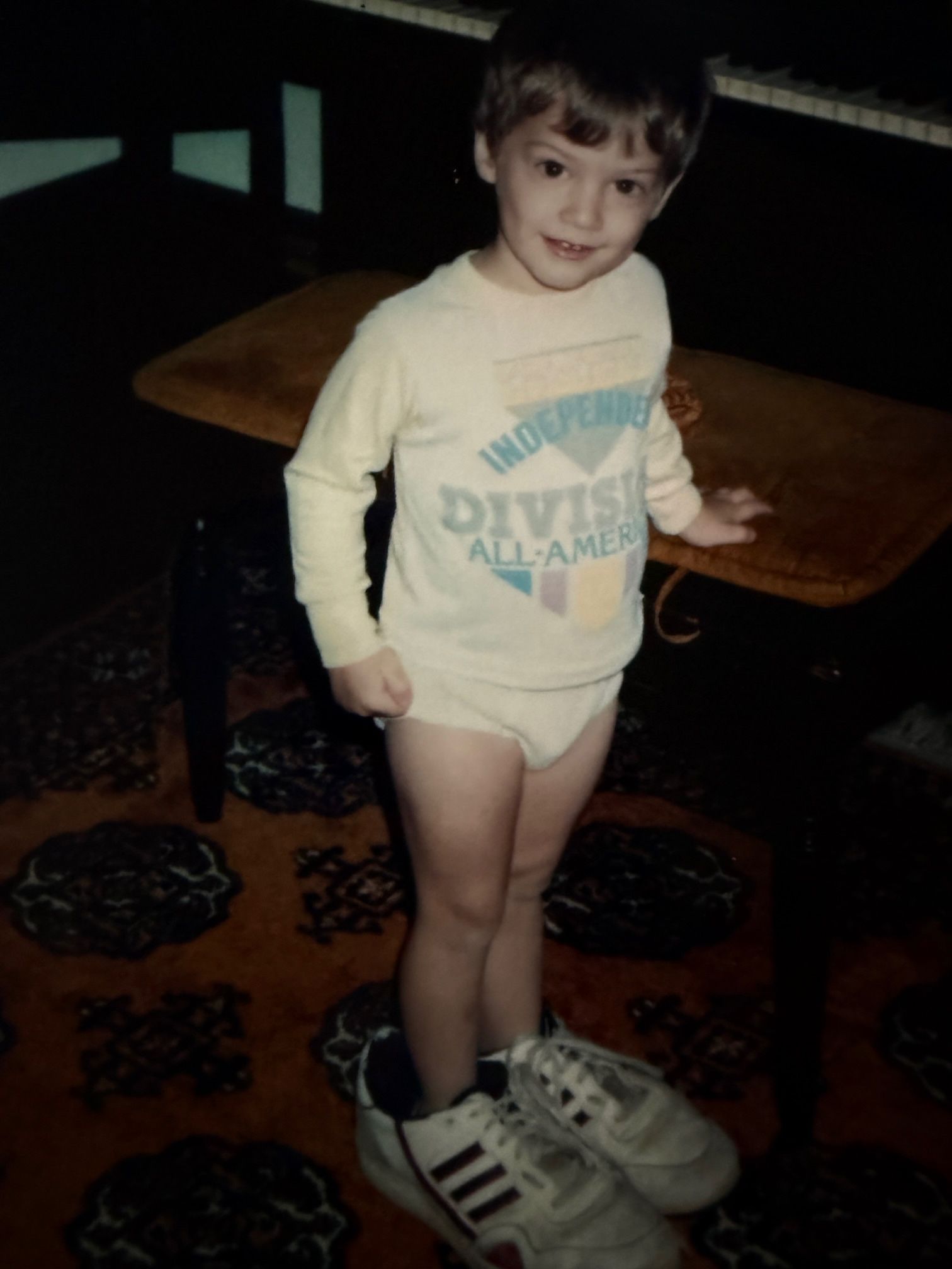 Boy wearing a large pair of sneakers, standing near a table and a piano. He is smiling.