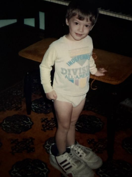 Boy wearing a large pair of sneakers, standing near a table and a piano. He is smiling.
