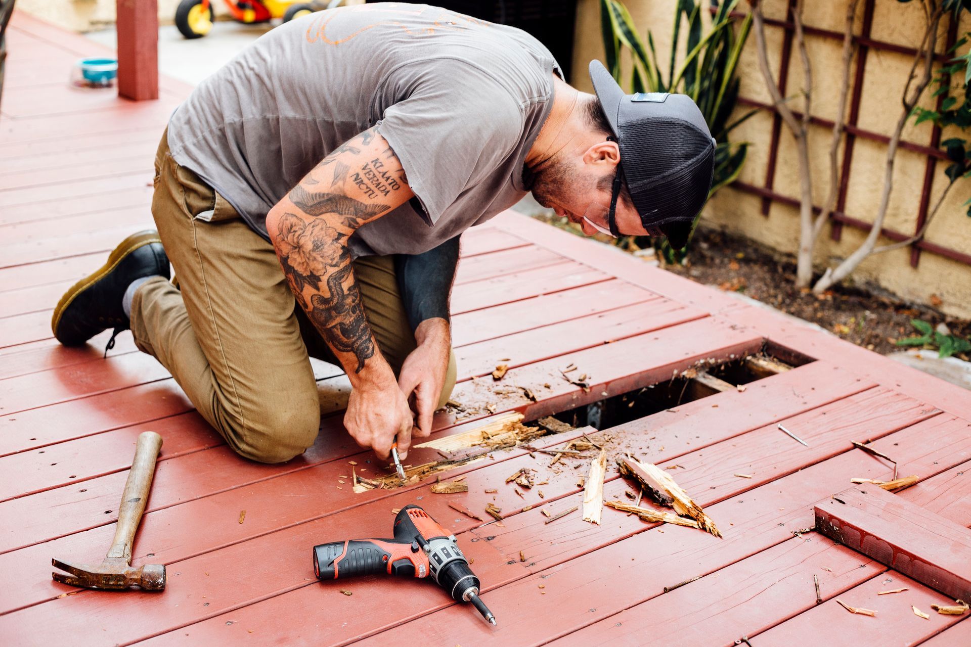 A man is kneeling down on a wooden deck with a drill and hammer.