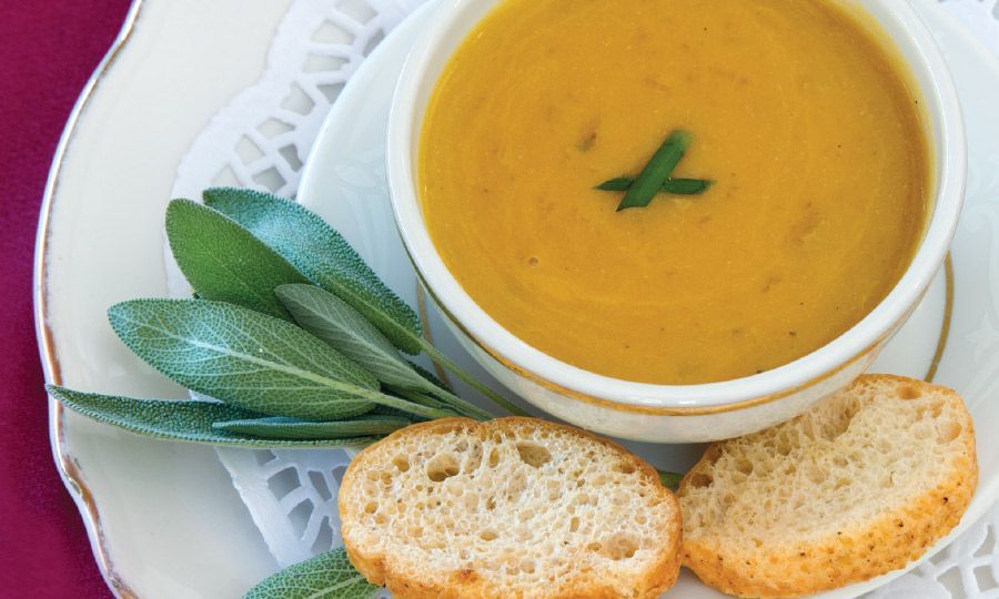Bowl of orange soup garnished with sage, served with bread on a white plate.