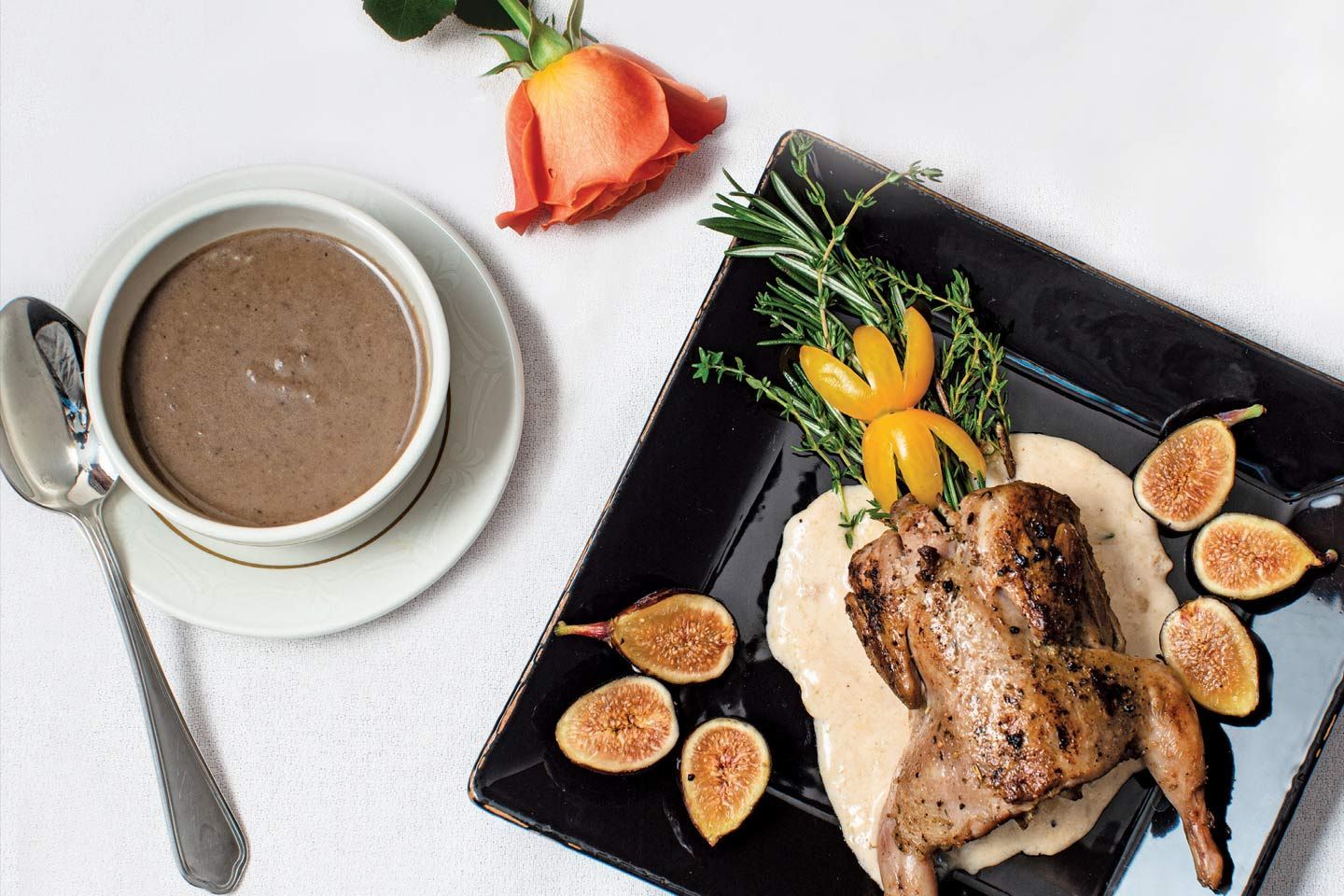 Gourmet meal on a white tablecloth: Roasted quail with figs, purée, herbs, and soup with a rose.