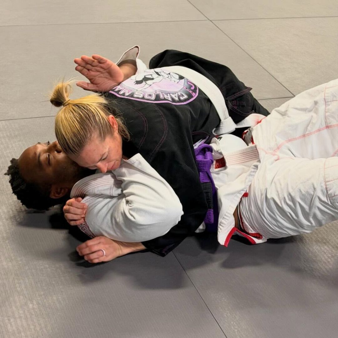 Two people practicing Brazilian Jiu-Jitsu on a mat; one in a black gi, one in a white gi, grappling.