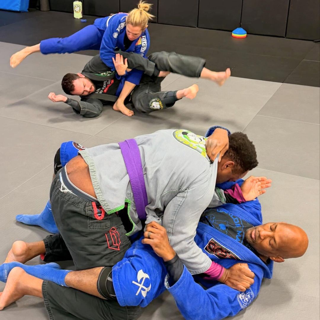 People practicing jiu-jitsu on mats, wearing gis in blue, black, and gray. Active grappling, gym setting.