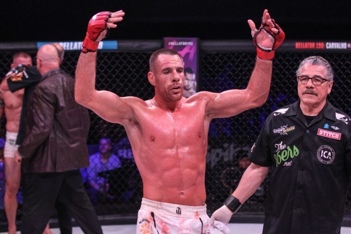 MMA fighter raises arms in victory, wearing gloves and shorts, in a brightly lit arena.