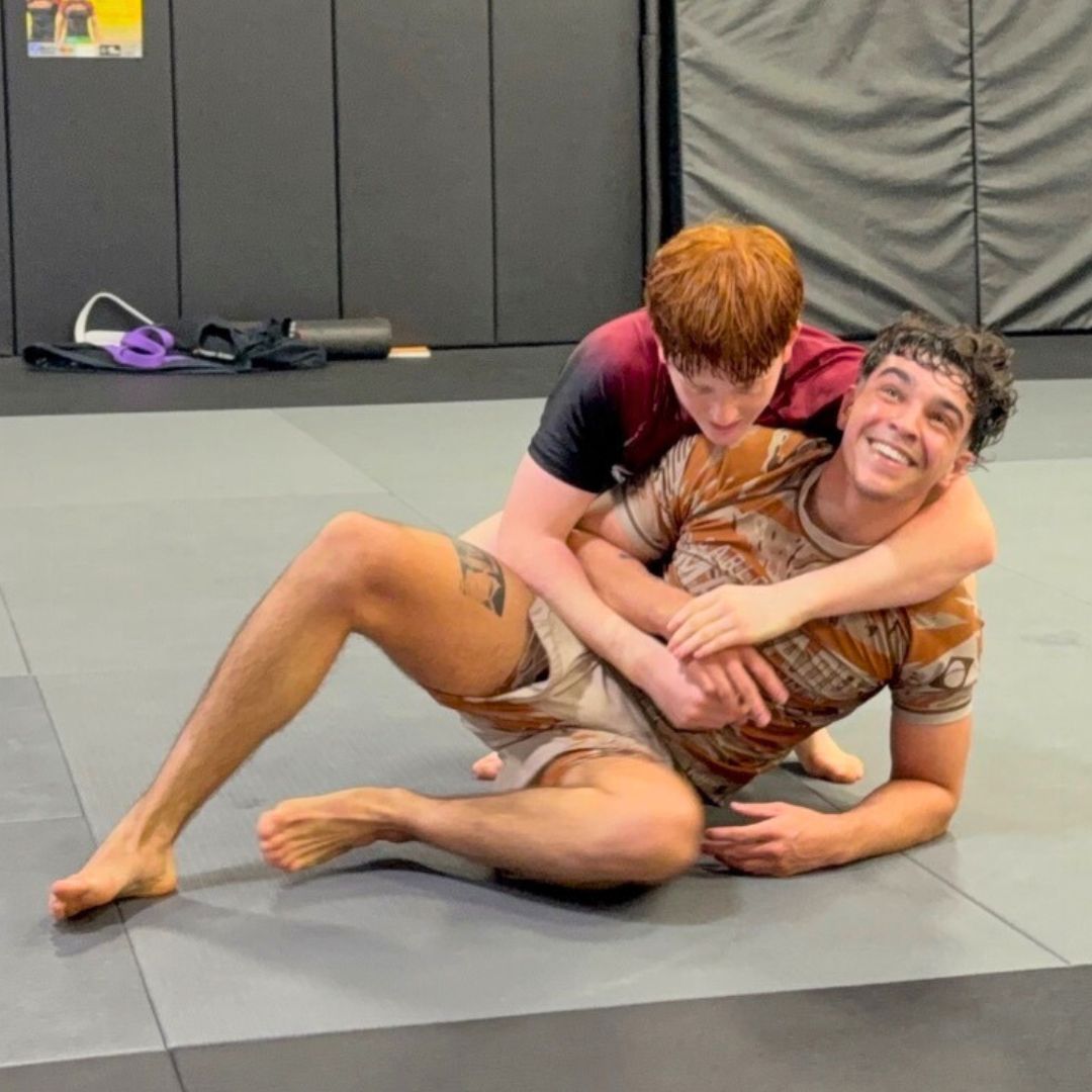 Two men in grappling position on a mat. One embraces the other from behind, both smiling. Gym setting.